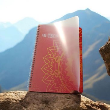 A spiral notebook with a translucent cover on a rock, mountains in the background.