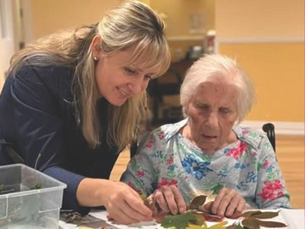A woman helps an elderly lady with a leaf craft activity indoors.