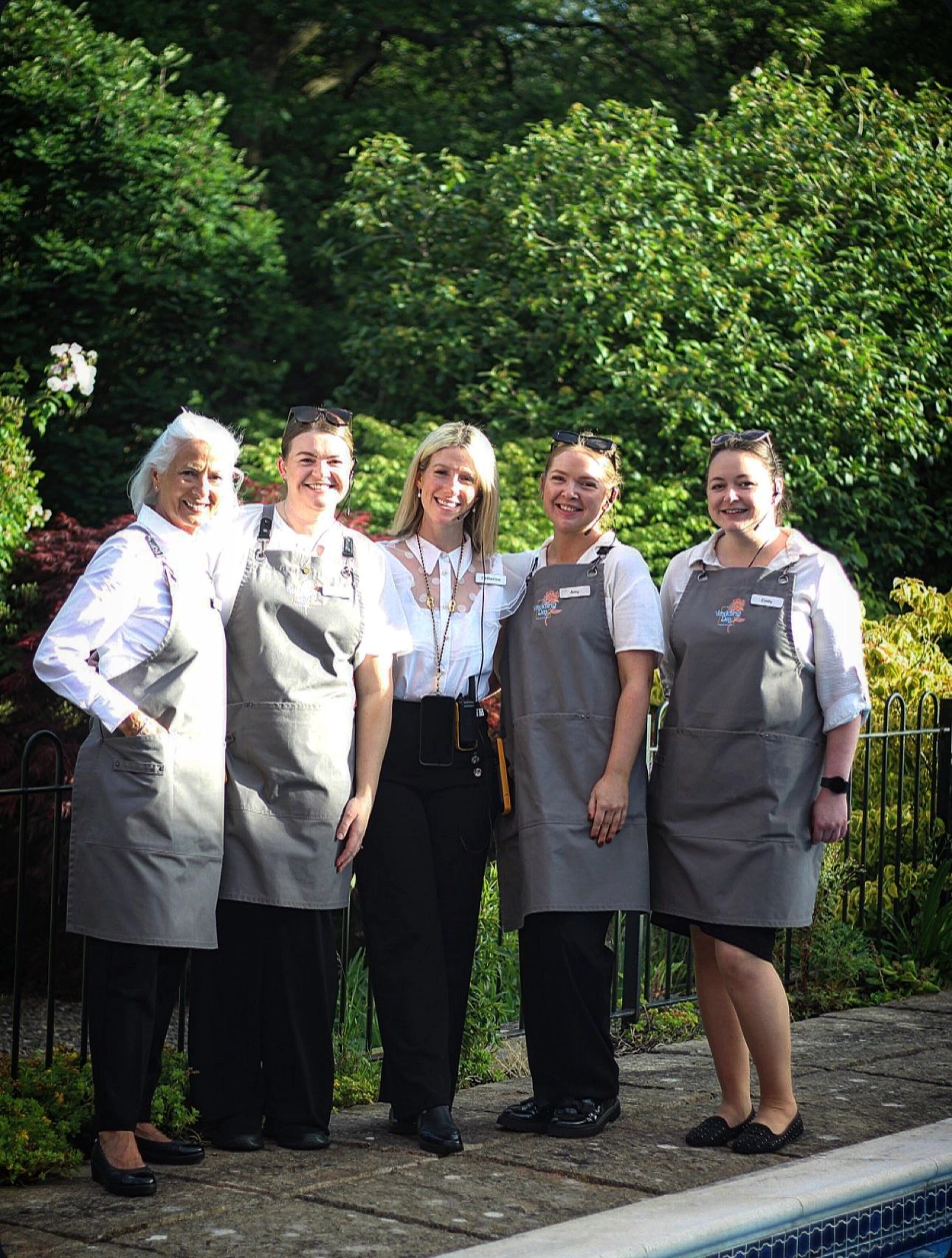 Five women smiling outdoors, four wearing matching aprons by a pool.