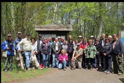 Group of people posing outdoors in front of a wooden shelter surrounded by trees.
