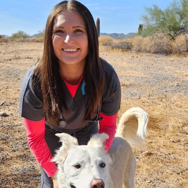 Smiling woman in scrubs with a white dog in a sunny desert landscape.