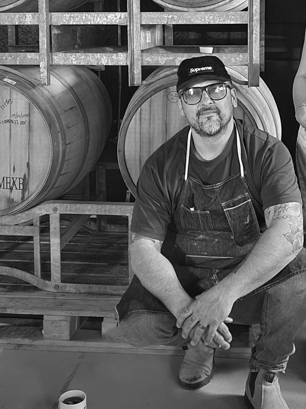 Man in apron sitting in front of wooden barrels.