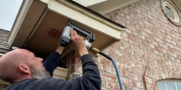 A man uses a nail gun to fix a wooden panel under a roof.