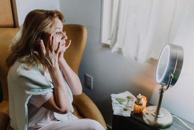 Woman sitting in a chair applying skincare in front of a mirror.