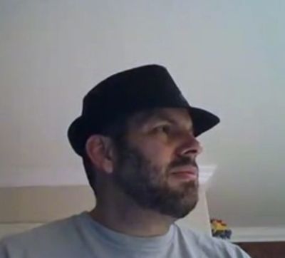 Man with a beard wearing a black fedora hat looking to the side indoors.