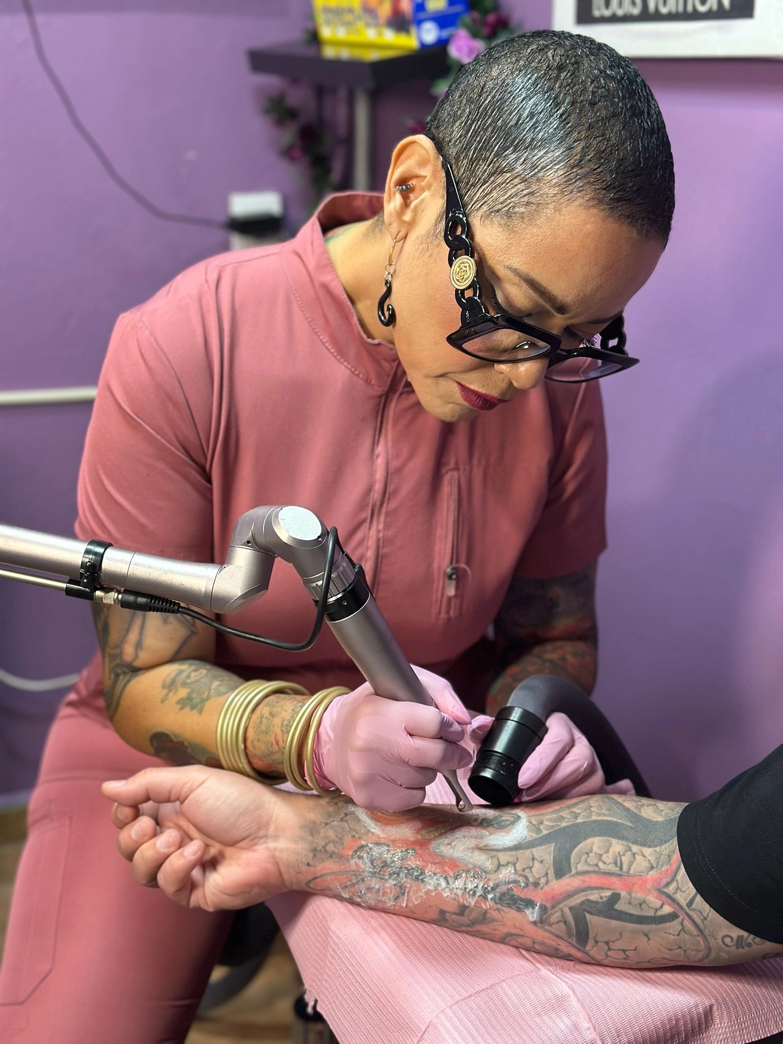 Smiling woman with blonde hair and tattoos holding a tattoo laser removal device. sacred body by jan