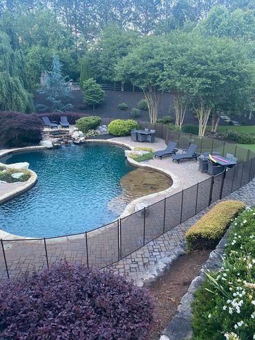 A backyard pool area with lounge chairs, tables, and a safety fence surrounded by lush greenery.