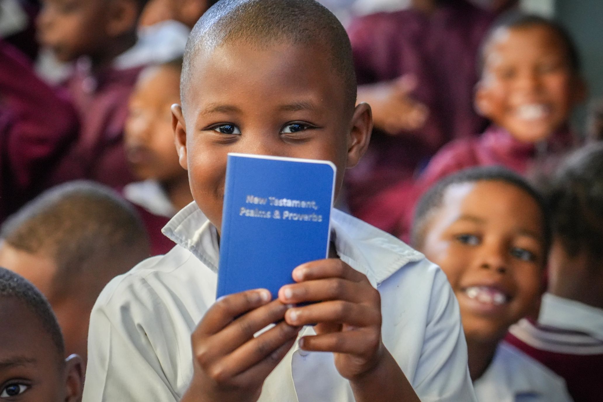 A child is holding a Gideon Bible and smiling. In the background are his classmates, also smiling.