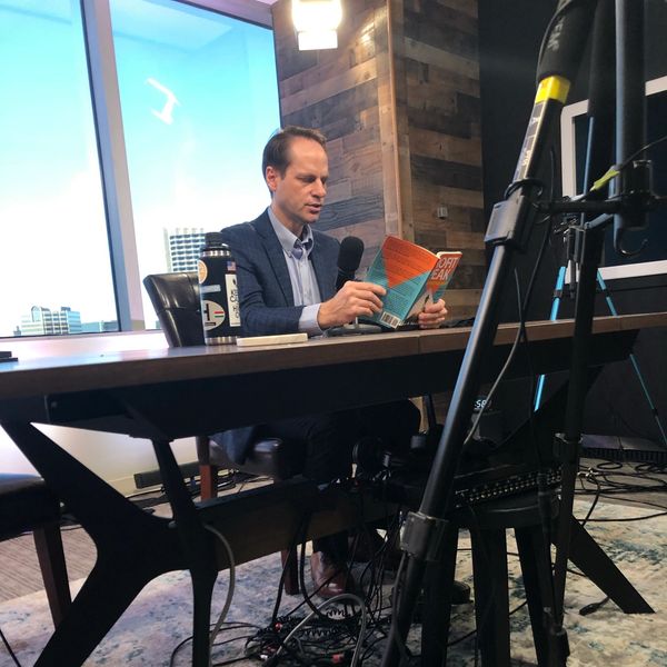 Man sitting at desk reading a book and holding a microphone.