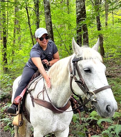 Woman in gray sportswear riding a white horse in a lush forest.