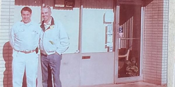 Two men standing outside a building entrance on a sunny day.