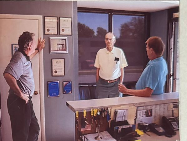 Three men engaged in conversation in an office lobby with framed certificates and a reception desk.