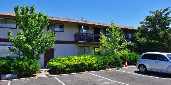 Apartment building with parking lot and green trees under clear blue sky.