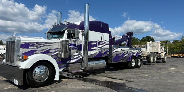 A purple and white tow truck with flame decals under a blue sky.