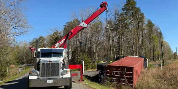 A crane truck lifting a tipped-over red shipping container on a rural road.