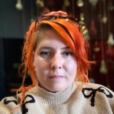 Femme aux cheveux roux portant un pull beige et des lunettes sur la tête.