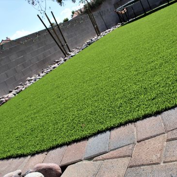 Large backyard with artificial grass, edged with pavers, and the back wall lined with river rock.