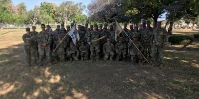 Group of soldiers in camouflage uniforms posing outdoors with flags.