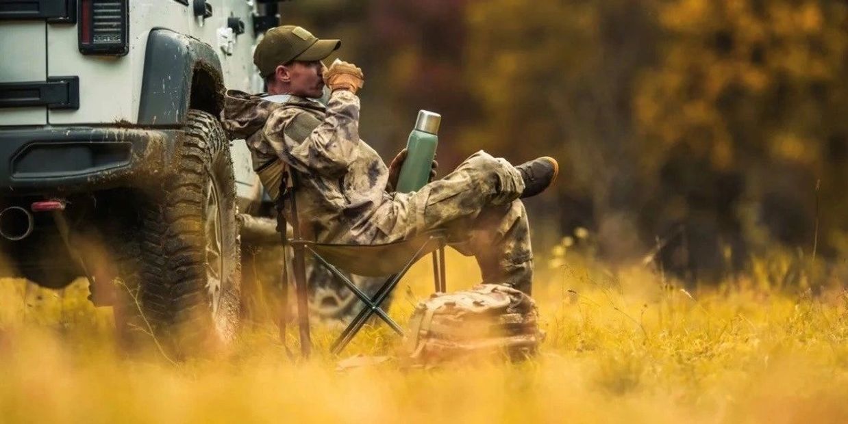 A person in camouflage drinks from a cup while sitting beside a vehicle in a field.