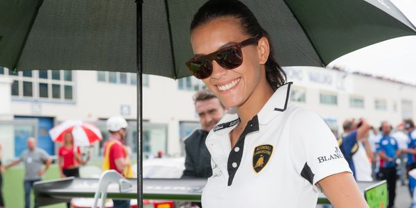 Smiling woman in sunglasses and white Lamborghini polo shirt under an umbrella at a racing event.