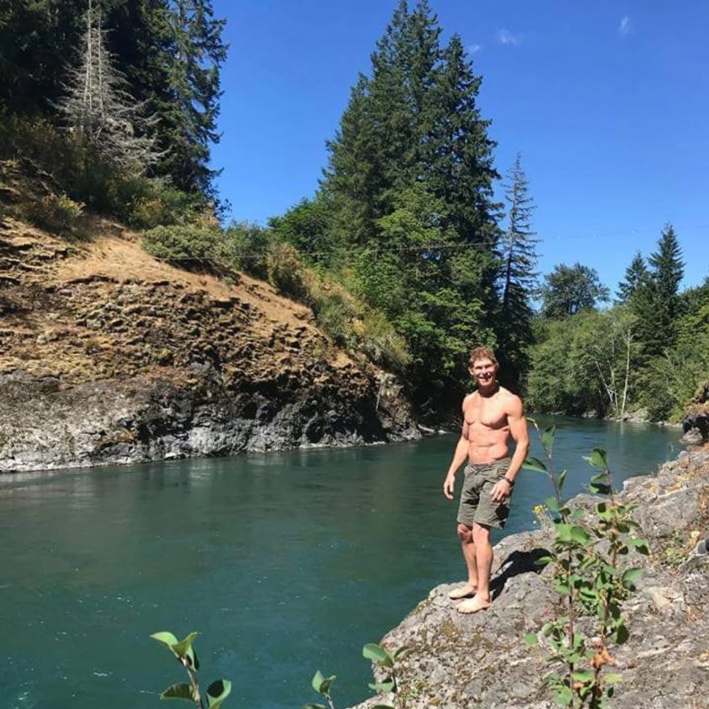 Shirtless man standing on rocky riverbank surrounded by trees.
