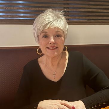 Smiling elderly woman with white hair and gold hoop earrings sitting at a table.