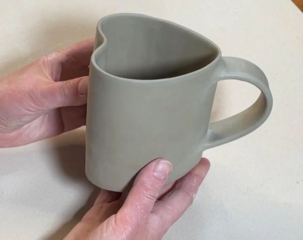 Hands holding a uniquely shaped heart-top ceramic mug.