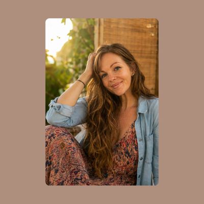 A smiling woman with long hair sits casually, bathed in warm sunlight.