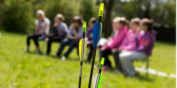 Three arrows stuck into a target outdoors, kids watching.
