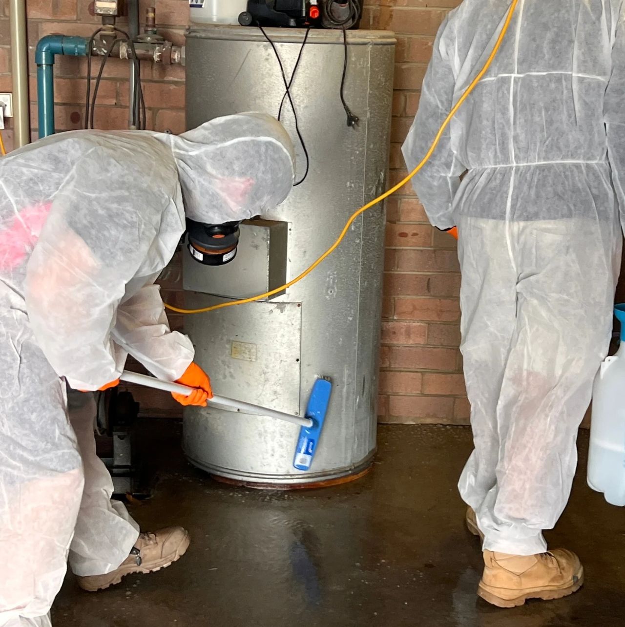 Restoration workers in protective suits cleaning water damage around hot water tank. Restoration workers in protective suits cleaning water damage around hot water tank.