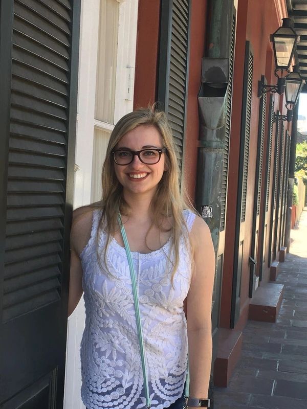 Smiling woman in a white lace top standing by a shuttered window on a sunny street.