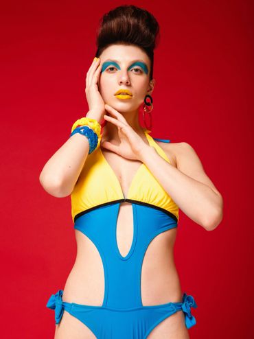 Model in vibrant blue and yellow swimsuit with bold makeup against a red background.