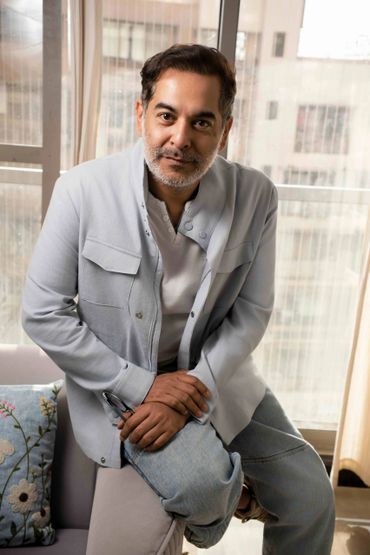 Man posing indoors wearing a light grey jacket and jeans.
