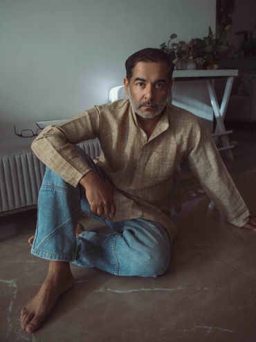 Man sitting barefoot on the floor in casual attire with a contemplative expression.