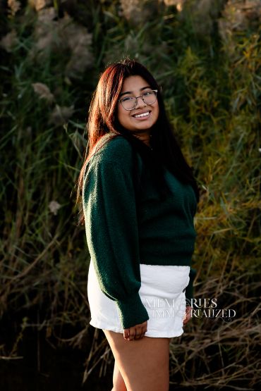 Young woman smiling outdoors in a green sweater and white skirt.