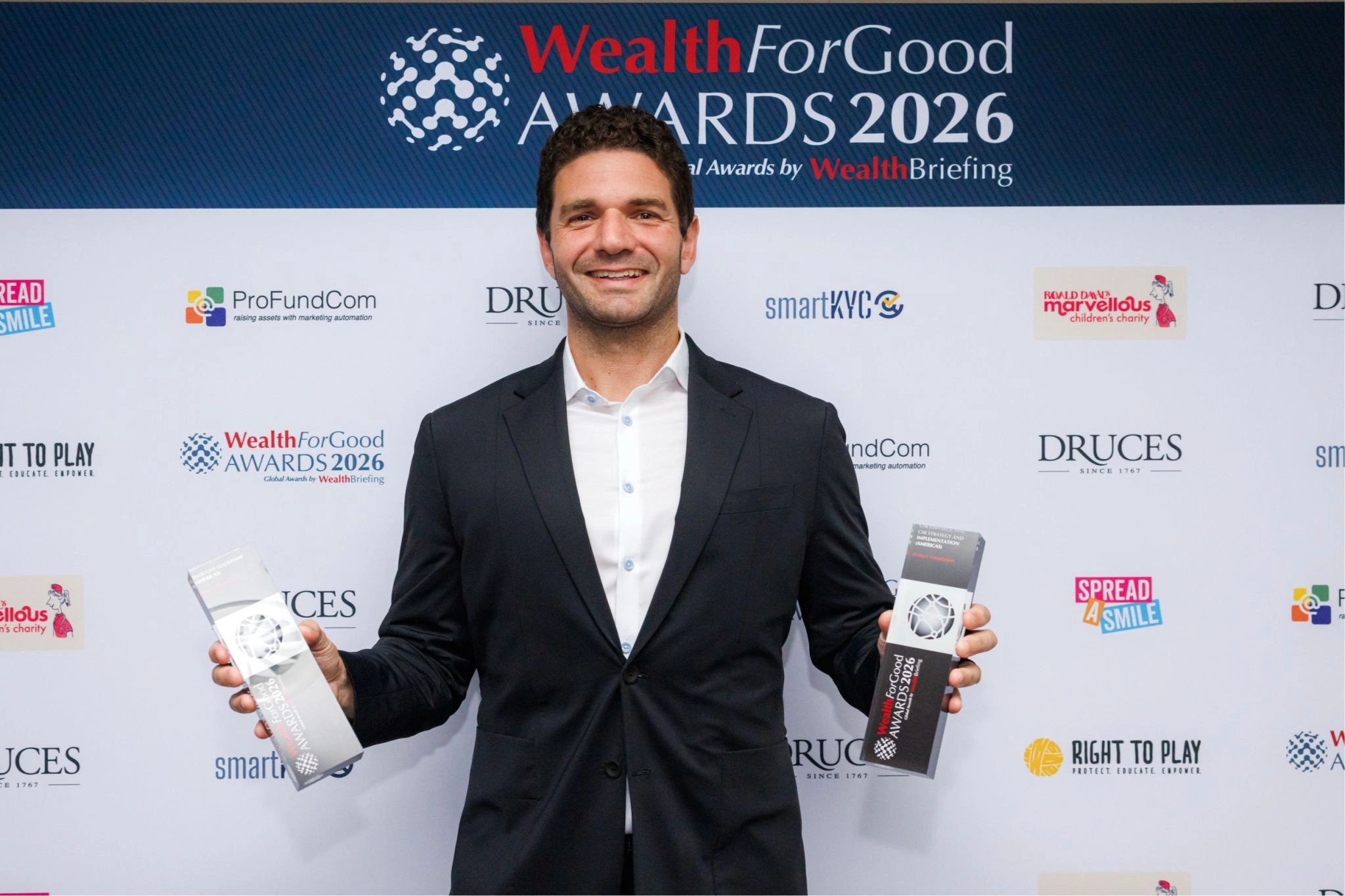 Smiling man in a suit holding two Wealth For Good Awards 2026 trophies.