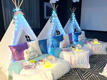 A dark room set up for a sleepover party. Three white teepee tents are decorated with fairy lights a