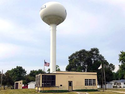 Tall water tower behind a small beige building with flags in a rural area.