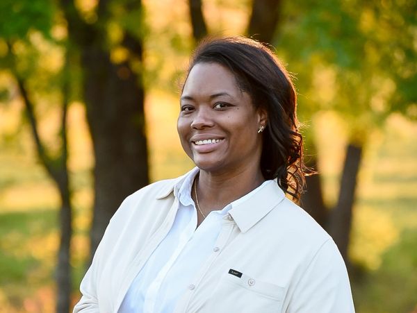 Smiling woman in white shirt outdoors with trees in the background.