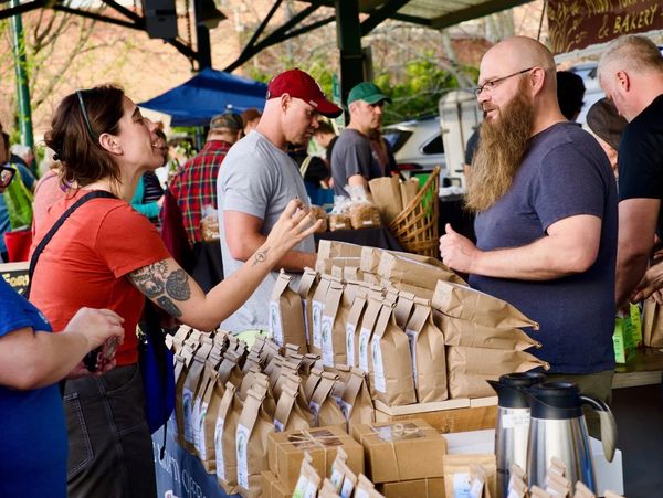 People interact at an outdoor market stall selling packaged goods and coffee.