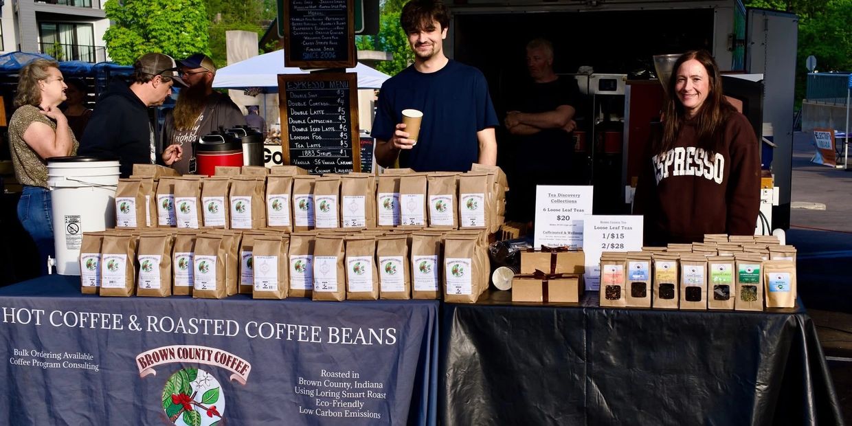 Two vendors at a market stall selling coffee and tea with many brown bags displayed.