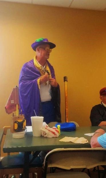 Man dressed in a purple cape and hat speaks to a seated group indoors.