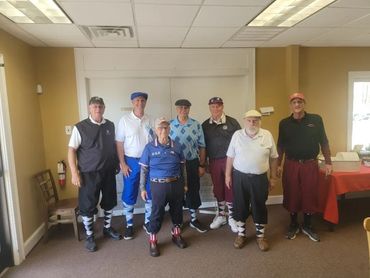 Seven men in golf attire posing indoors, each wearing knee-high golf gaiters.