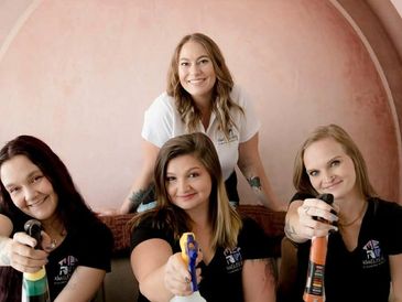 Four women smiling, holding spray bottles, posing in a cozy setting.