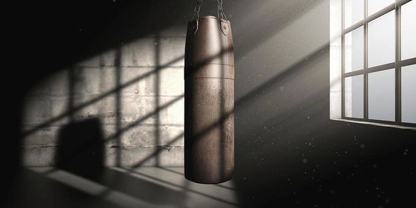 A punching bag hangs in a dimly lit gym with sunlight streaming through a window.