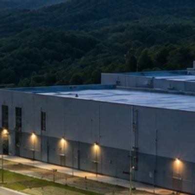 Exterior of a large industrial building with lights on at dusk.