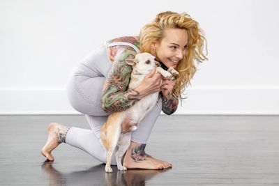 Smiling tattooed woman crouches and hugs a small dog indoors.
