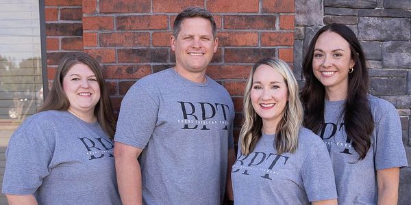 Four adults wearing matching gray BDT t-shirts smiling by a brick wall.