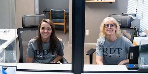 Two women in matching gray RDT shirts sitting at office desks separated by a glass partition.