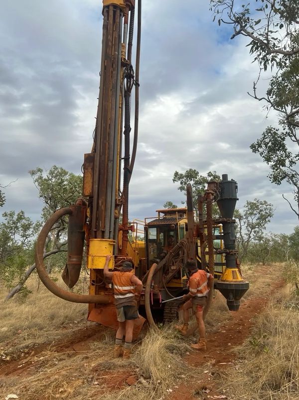 A yellow drilling machine operating in a forested area under a cloudy sky.